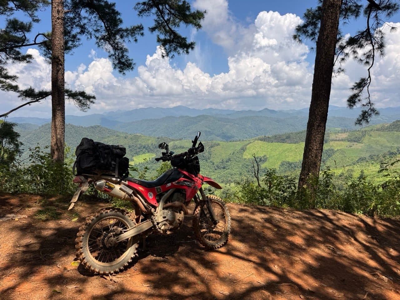 A red Honda CRF250L parked at a pine-framed viewpoint on the Tail of the Elephant, with forested ridges running out behind